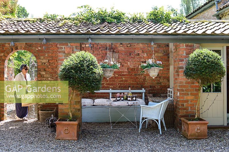 Covered seating area with wooden painted bench and table under overhang, standard trees in pots and hanging baskets 