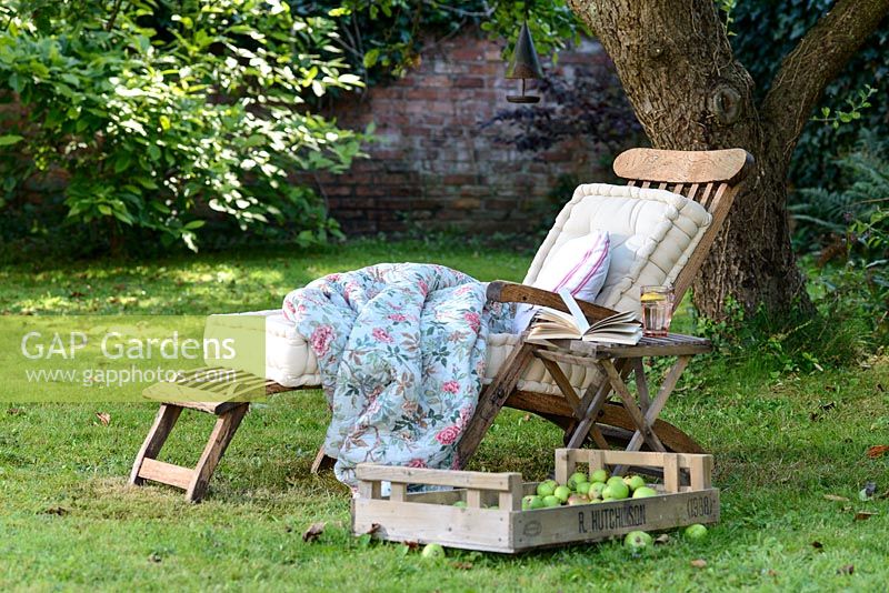 Wooden deckchair lounger with cushions and blanket on a lawn in the shade under a tree 