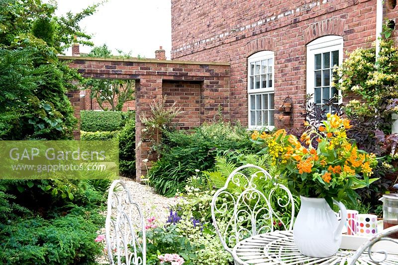 Small courtyard with decorative white metal furniture and vase with flowers, foliage bed against brick house and wall