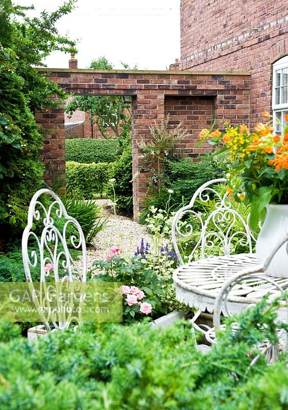 Small courtyard with decorative white metal furniture and vase with flowers, view towards brick house and wall