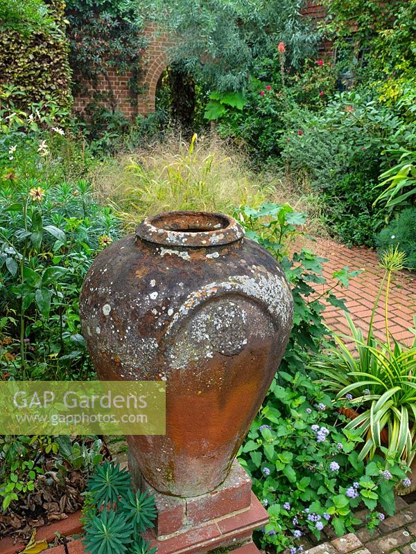 Clay urn on plinth at corner of bed with brick path beyond 