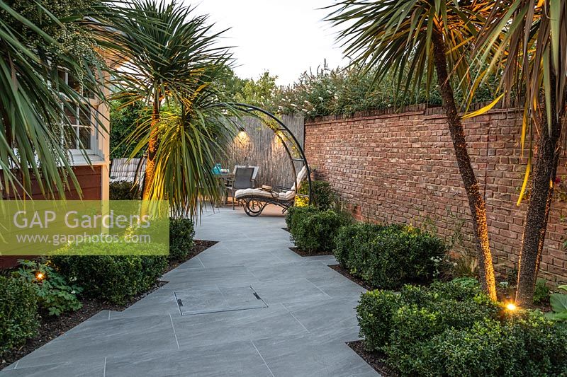 View along paved path with narrow beds with Cordyline illuminated with uplighters, beyond wall-mounted downlighters by seating