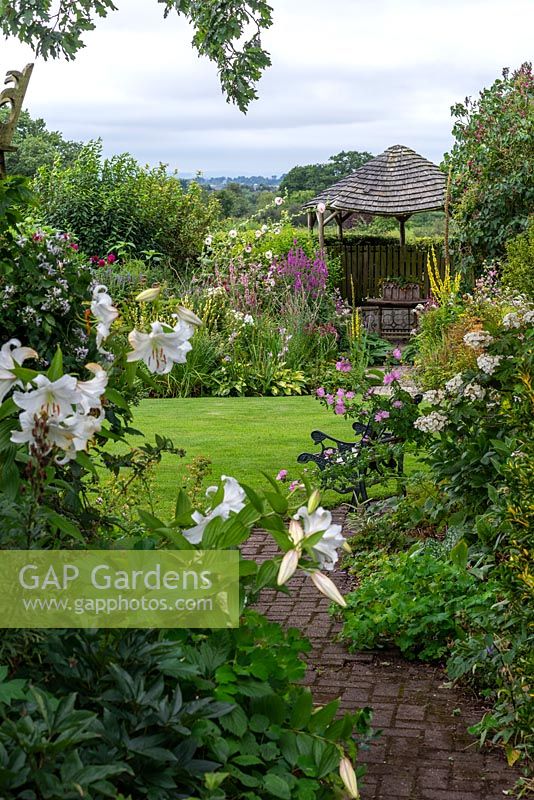 Views along a brick path to a wooden gazebo, flanked by borders of summer herbaceous perennials. Beyond, glimpses of Blackmore Vale.