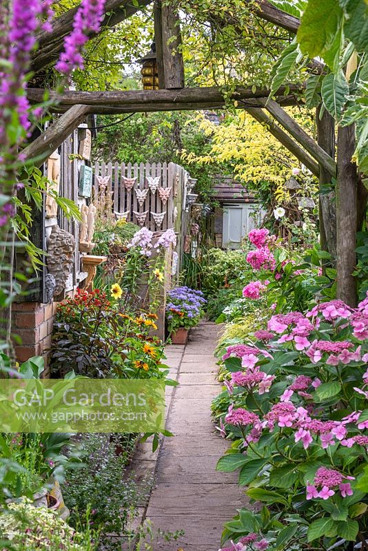 Rustic arch frames a view along a path, leading past pink hydrangeas and phlox. On left, hot coloured dahlias and rudbeckias.