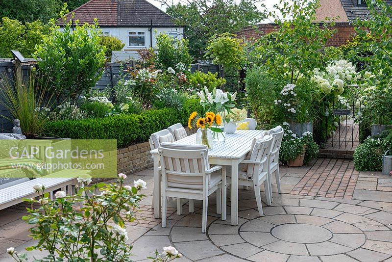 Table and chairs in paved dining area in small town garden, with white-themed borders. 