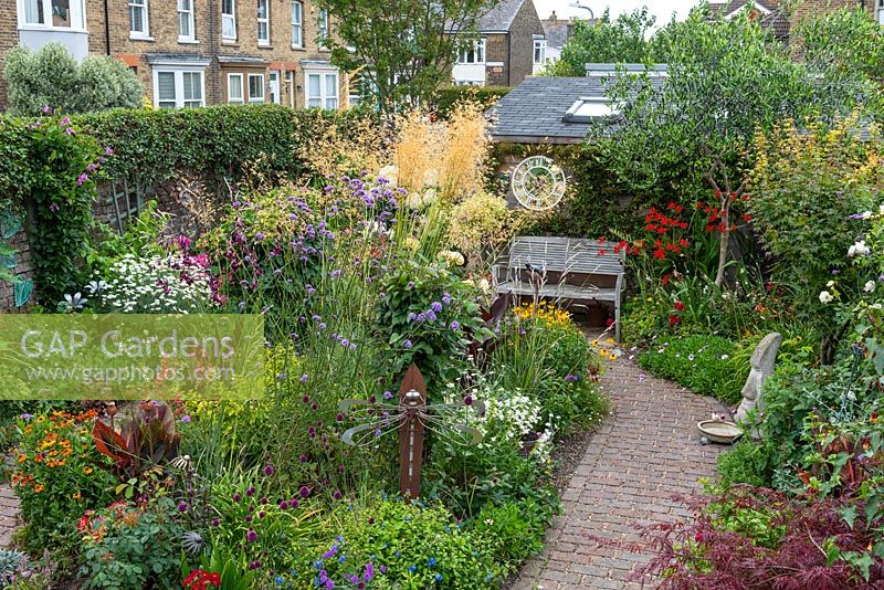 In a 7.5m x 14m back garden, a brick path in shape of big '6' separates side borders and rear shed from a central bed planted with Stipa gigantea, Verbena bonariensis, roses, dahlias, heleniums, alliums, penstemon, heucheras, hydrangeas and cannas.