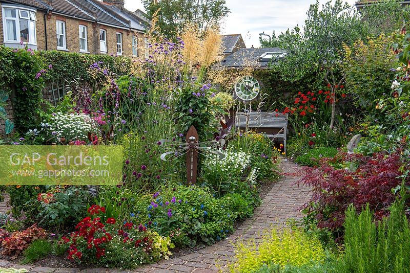 In a 7.5m x 14m back garden, a brick path separates side borders of herbs and acer from a central bed planted with Stipa gigantea, Verbena bonariensis, roses, dahlias, heleniums, alliums, penstemon, heucheras and cannas.