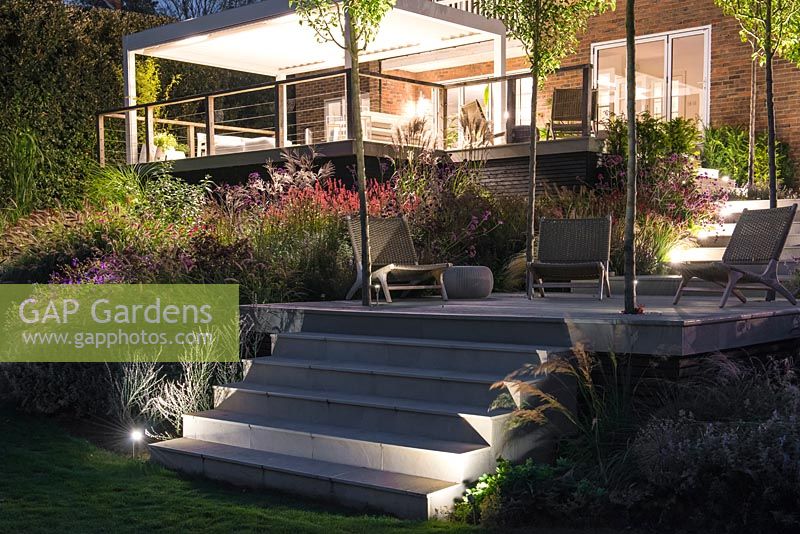 Contemporary decks and steps lit at night, illuminating surrounding borders of ornamental grasses and flowering perennials.