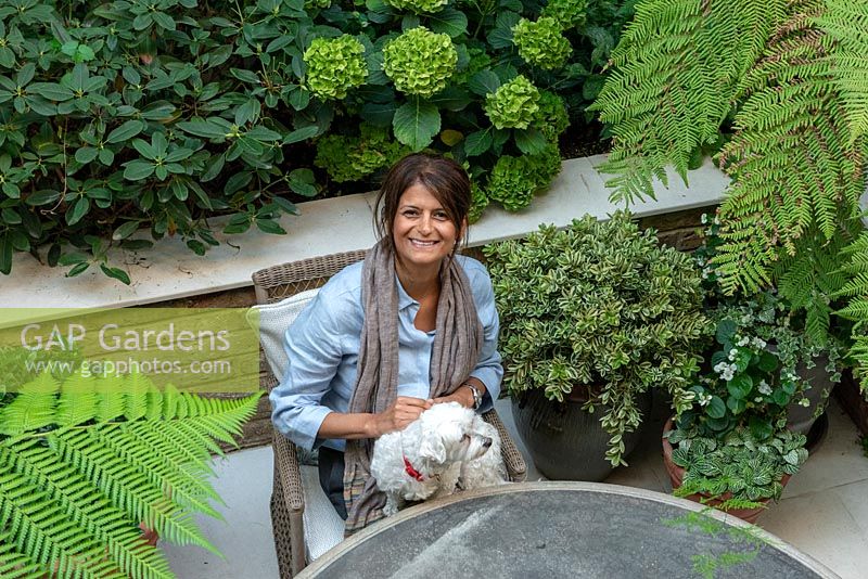 Woman sitting in her small courtyard garden with pet dog on her lap, surrounded by green foliage plants