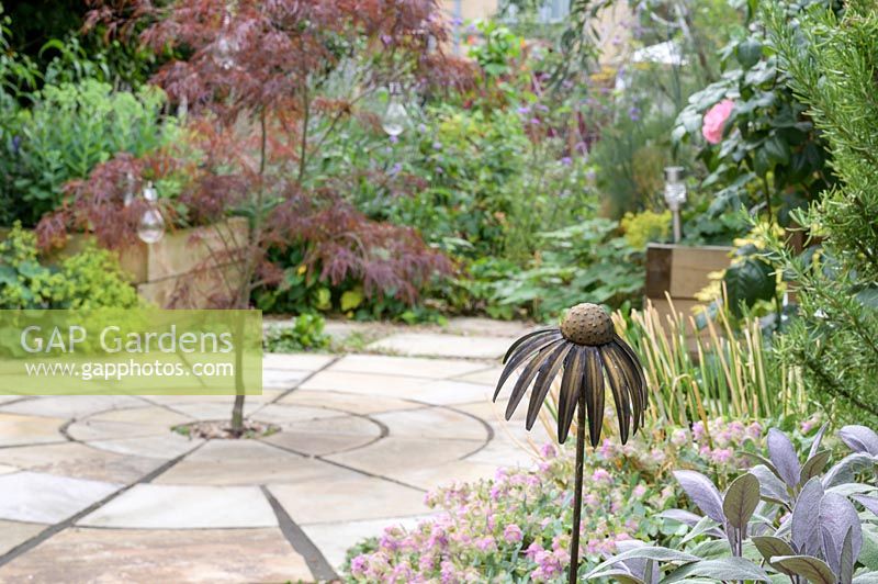 Metal flower head in a border, beyond circular paving with centrally planted Acer - Maple