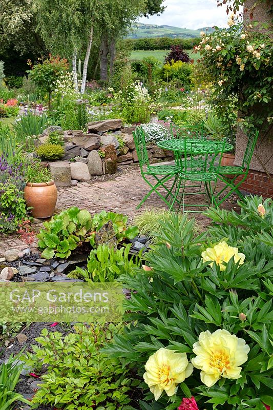 View across small patio and rockery to flower beds and countryside, in foreground Paeonia 'Bartzella' 