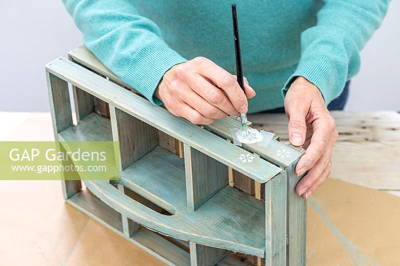 Woman stencilling small snowflakes on to the side of the wooden box using white paint. 
