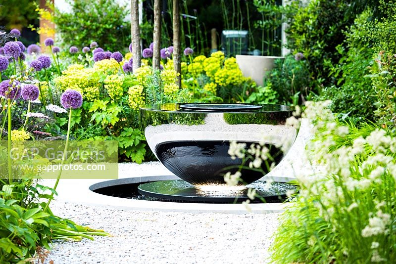 View of David Harber water feature surrounded by Allium 'Globemaster', Luzula nivea and Euphorbia characias subsp. wulfenii. 