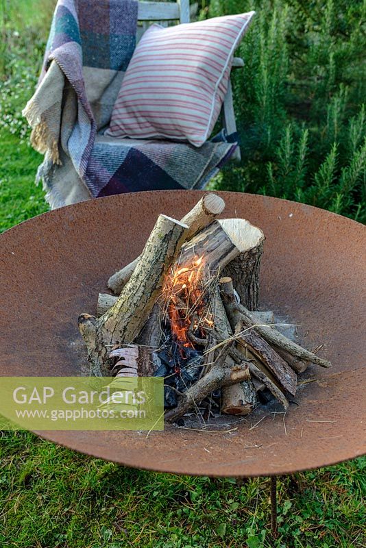 A lit fire bowl made from rusted corten steel, a wooden garden chair with a wool rug and a cushion by a large rosemary bush.