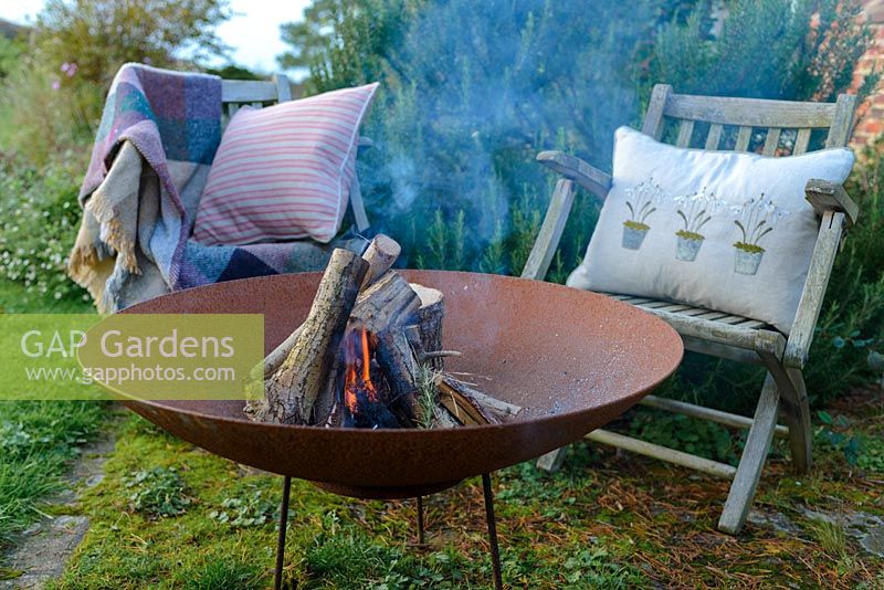 A lit fire bowl made from rusted corten steel,  wooden garden chairs with a wool rug and  ushions by a large rosemary bush.