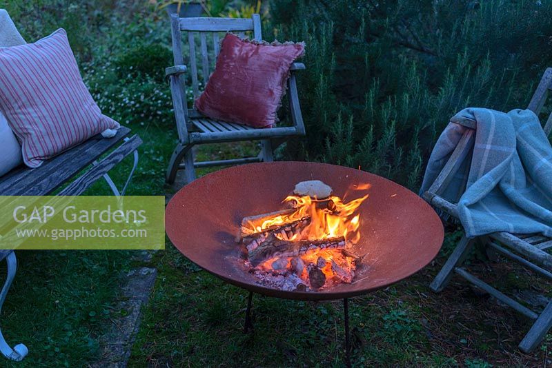 A lit fire bowl made from rusted corten steel,  wooden garden chairs and a bench with wool rugs and  cushions by a large rosemary bush.
