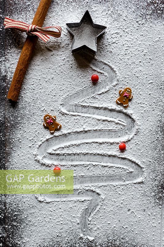 A Christmas tree drawn in icing sugar on a wooden chopping board with a star cutter, ginger bread men, red chocolate buttons and a cinnamon stick tied with ribbon.