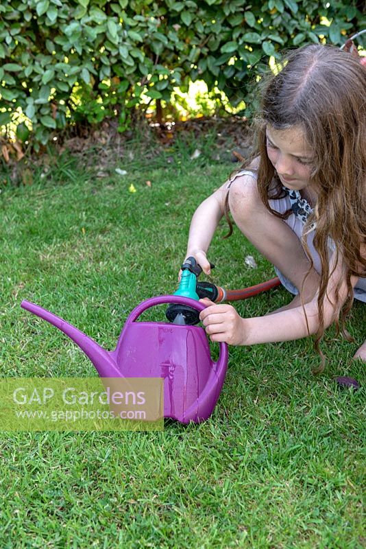 Girl filling plastic watering can with water from the hose. 