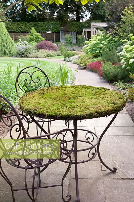 A shady garden feature of green moss growing on a cast iron metal round table with chairs on patio - July