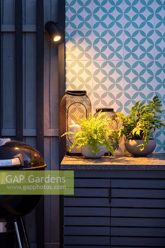 Illuminated worktop with ferns planted in small pots and lanterns by colorful wall in an outdoor kitchen at night.