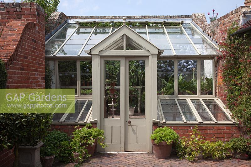 Small wooden greenhouse set in brick walls in courtyard garden, with cold frames with tender plants growing inside. 