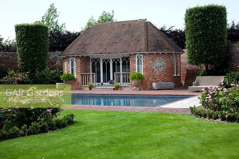 Swimming pool and pool house in walled garden with brick paving, edged with large topiary columns and  herbaceous borders filled with pink roses.