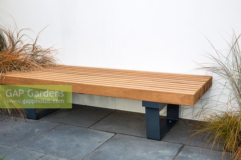 A modern wooden garden bench with metal frame legs on a dark stone paved patio. The Low Line Garden, RHS Malvern Spring Festival, 2016. Design: Jamie Langlands. 