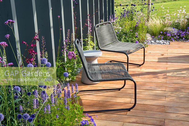 Metal framed black chairs on all weather mandioqueira wood decking with grey painted wooden vertical fence boards.  Business and Pleasure Garden, RHS Tatton Park Flower Show, 2017. Designer: Jake Curley