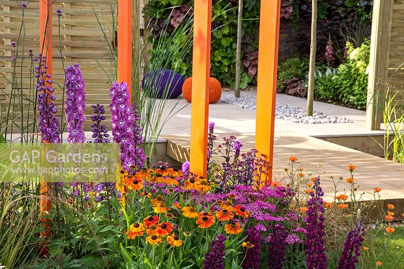 Flowerbeds filled with colourful flowering plants including Helenium, Delphinium,  Achillea millefolium, Geum 'Totally Tangerine' and Lupins in borders with painted orange screen dividers - For The Love Of It garden. Tatton Flower Show 2017 Designer: Pip Probert 