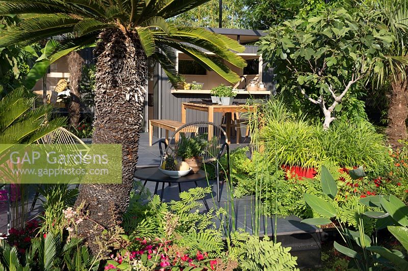 View towards outdoors living space metal outdoor kitchen bar summerhouse on a dark graphite porcelain stone patio seating area with wooden bench and table - planting of tropical style plants Busy Lizzie 'Imara' with ferns, grasses, palm and fig trees, horsetail and bamboo plants. RHS Hampton Court Palace Flower show July 2018 B and Q's Bursting Busy Lizzie Garden - Designer: Matthew Childs - Sponsored by B and Q 