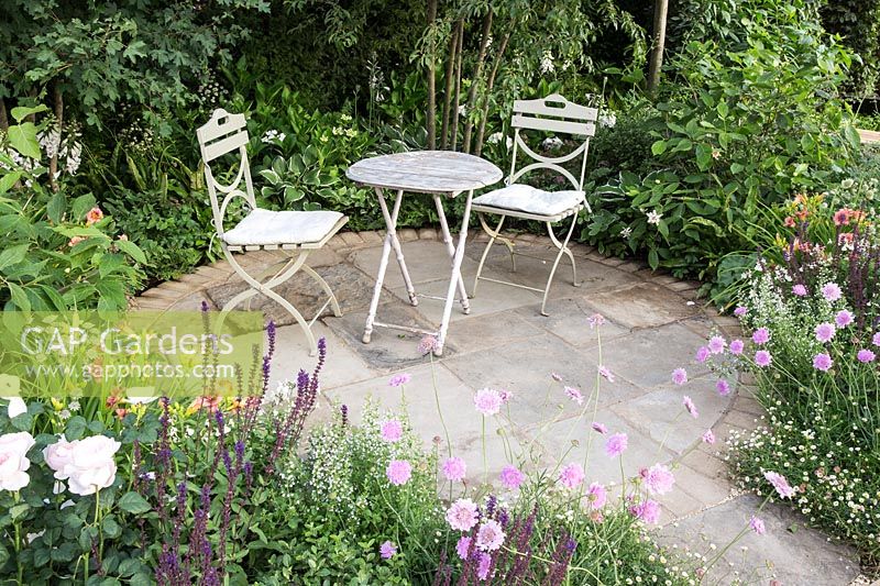 Cottage garden with a round patio with small table and chairs, surrounded by beds with: Scabiosa incisa 'Kudo', Salvia nemorosa, roses, hostas and Erigeron karvinskianus. BALI Best of Both Worlds Garden