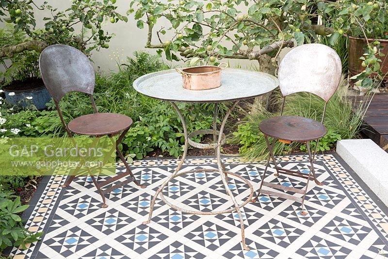 Metal table and chairs on a tiled mosaic patio, behind bed with Malus - Apple - trees and underplanting.The Style and Design Garden, sponsors CED Stone, London Mosaic, Garden Brocante Online