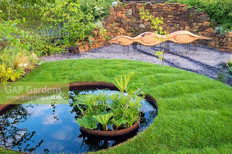 Small round pond water feature planted with Gunnera manicata and Papyrus- carved oak wood bench on gravel seating area - small undulating grass lawn. Grace  and  Dignity garden. RHS Malvern Spring Festival May 2019 -  Designer Lucie Giselle Ponsford 