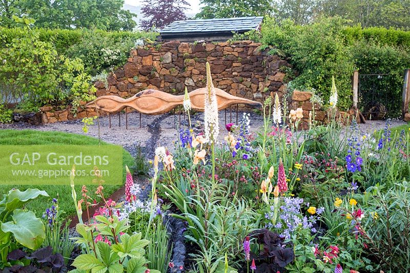 Small flower bed planted with Irises, Eremurus, delphiniums, lupins, Primula viallii - carved oak wood bench on gravel seating area - small grass lawn. Grace and Dignity garden. RHS Malvern Spring Festival May 2019 -  Designer Lucie Giselle Ponsford - 