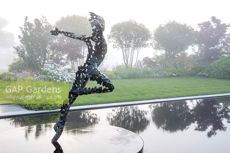 Reflective black pond water feature with sculpture 'Zephyr' by Simon Gudgeon on a misty May spring day - small grass lawn with mixed herbaceous planting. Garden of Quiet Contemplation. RHS Malvern Spring Festival 2019 - Designer Peter Dowle - Leaf Creative - 