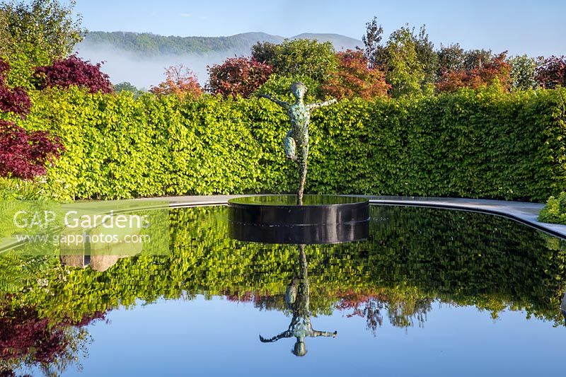 Reflective black pond water feature with sculpture 'Zephyr' by Simon Gudgeon on a misty May spring day surrounded by garden hedge and acers. Garden of Quiet Contemplation garden.  RHS Malvern Spring Festival May 2019 - Designer Peter Dowle - Leaf Creative 