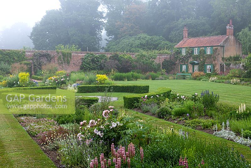 An old cottage overlooks the walled Spider Garden at Hoveton Hall. A grassy path separates herbaceous borders, a box circle halfway along.