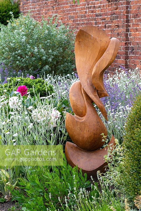 Wooden sculpture by Peter Leadbeater in a herbaceous border of Lychnis coronaria 'Alba', catmint and Peonies.
