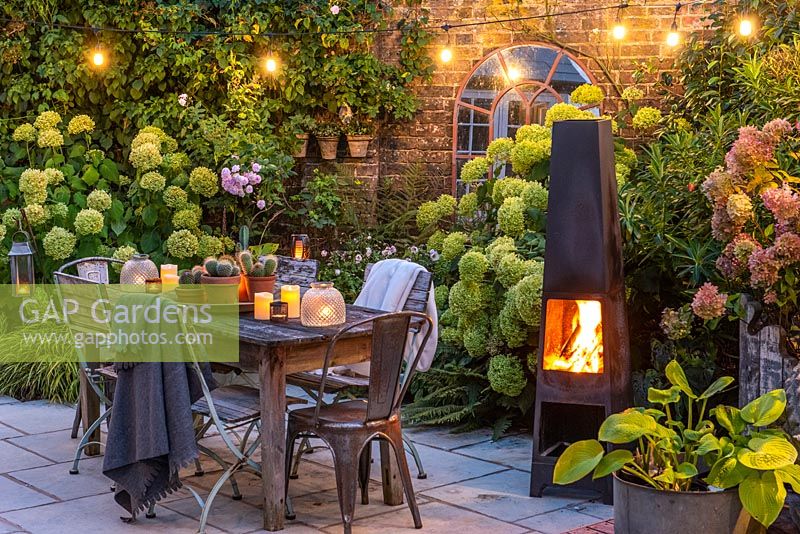 A dining area in a courtyard garden lit by a string of bulbs and candles, heated by a modern outdoor chimenea. Planting behin includes Hydrangea 'Limelight', Rosa 'Blush Noisette', Anemone hybrida 'Elfin Swan' and ferns.
