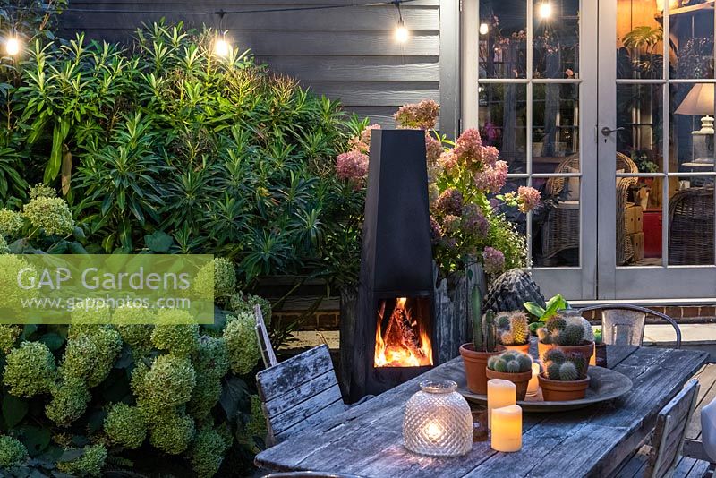 A dining area in a courtyard garden lit by a string of bulbs and candles, heated by a modern outdoor chiminea. Planting behind includes Hydrangea 'Limelight', Rosa 'Blush Noisette' and ferns.