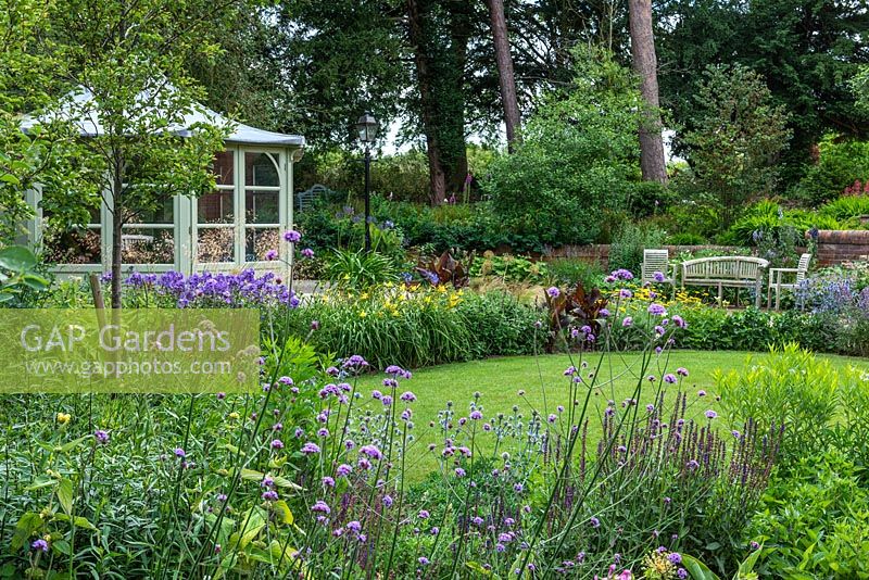 Borders of Sea holly and Verbena bonariensis, an oval lawn edged in herbaceous perennials. Beyond, a terrace with bench and chairs, overlooked by a summerhouse.