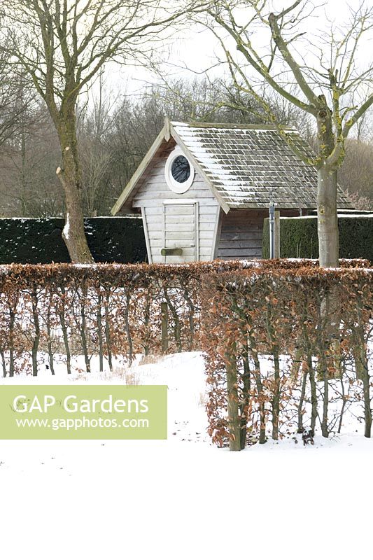 Gardenhouse with round window behind hedges in the snow.