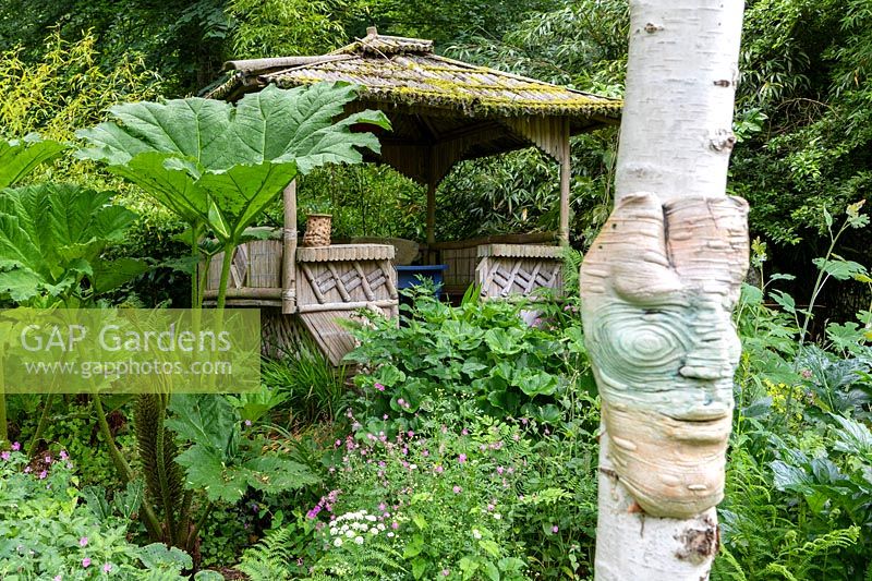 Ceramic face on tree trunk with view to summerhouse set in lush garden with bold foliage