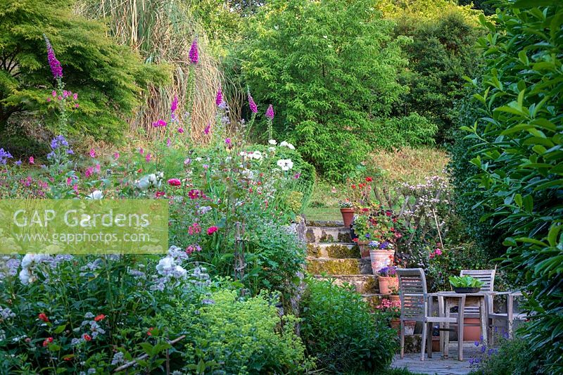 View along flower border to a patio area with wooden garden furniture, steps up to woods in country cottage garden