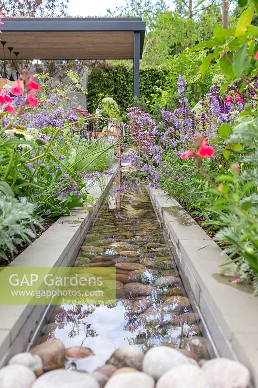 Looking down a contemporary rill water feature lined with large pebbles towards modern covered outdoor room. The Viking Cruises Lagom Garden - Hampton Court Flower Festival 2019