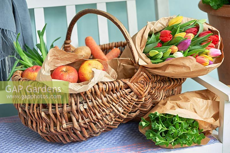 Basket with apples, carrots, spring onions, spearmint and wrapped tulips displayed on bench.
