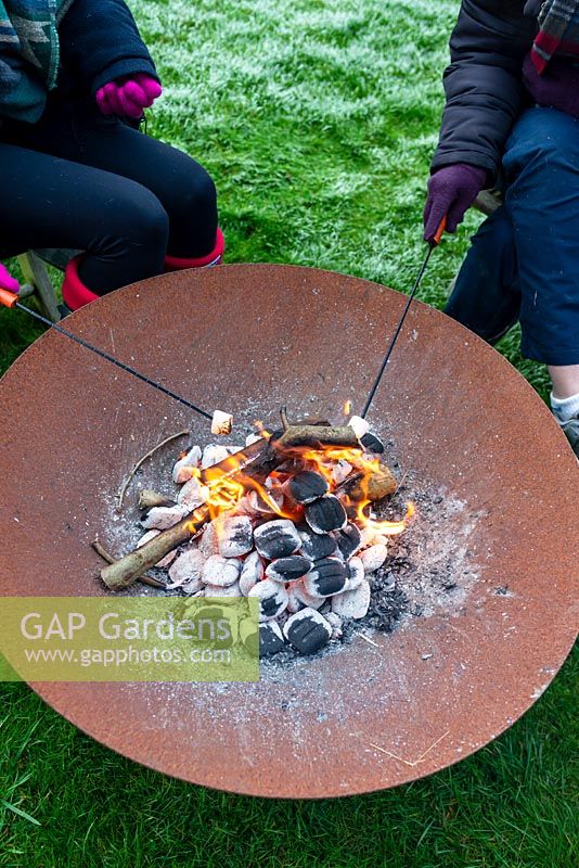 People wrapped in warm clothes, roasting marshmallows over a fire in a Corten Steel fire pit on a frosty winter day.