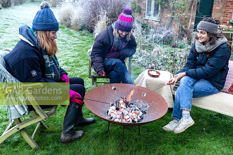 Women wrapped in warm clothes, roasting marshmallows over a fire in a Corten Steel fire pit on a frosty winter day.