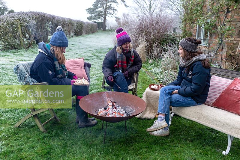 Women wrapped in warm clothes, roasting marshmallows over a fire in a Corten Steel fire pit on a frosty winter day. 