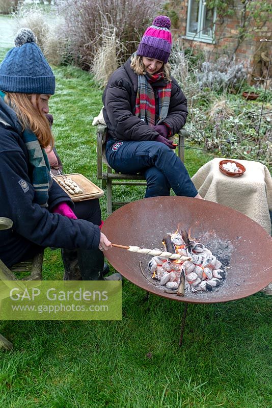 Women wrapped in warm clothes, cooking camp fire bread over a fire in a Corten Steel fire pit on a frosty winter day